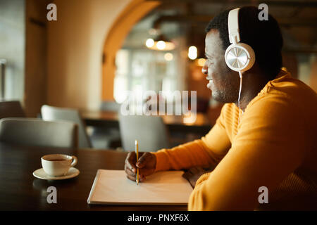 Young casual man with blank paper and pencil sitting by table and listening to audio training about sketching principles in headphones Stock Photo