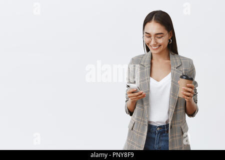 Girl checking messages, talking with friends, making new appointment. Portrait of confident busy female entrepreneur in glasses and jacket, holding cup of coffee and smartphone, using gadget Stock Photo
