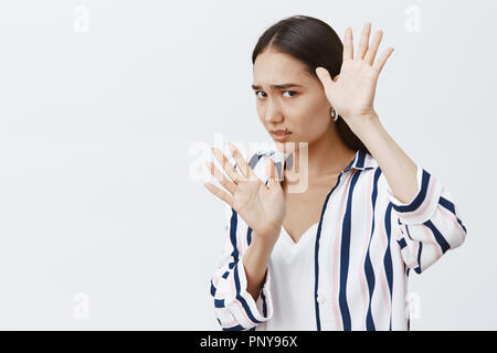 Victim of domestic violence. Portrait of scared timid woman in striped blouse, covering face with raised palms, defending from punch, being afraid of husband who beats wife over gray background Stock Photo