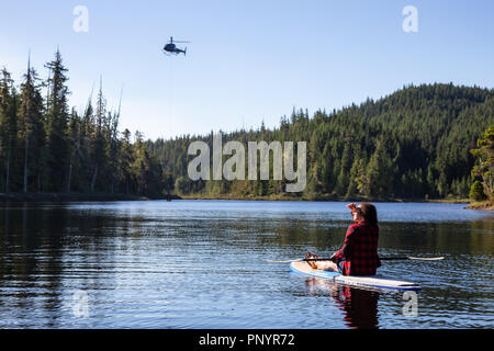 Northern Vancouver Island, British Columbia, Canada - August 17, 2018: Girl on a paddle board is watching an helicopter fighting BC Wild Forest Fires. Stock Photo