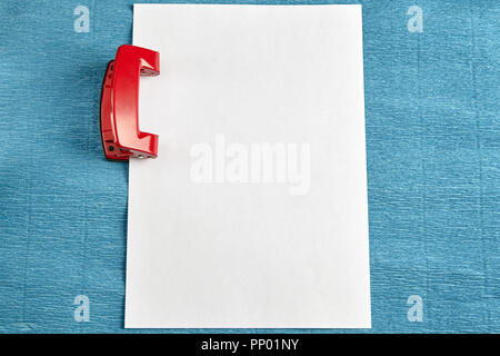 Red office punch with blank sheet of paper, blue background. Stock Photo