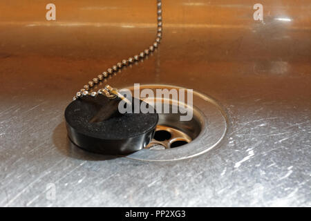 Black plug on beaded chain sits next to a plug hole in a stainless steel kitchen sink. Stock Photo