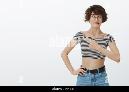 Portrait of unsure hesitating cute girlfriend in round glasses and striped cropped top, biting lip while thinking or making choice, looking and pointing at upper left corner, doubting Stock Photo