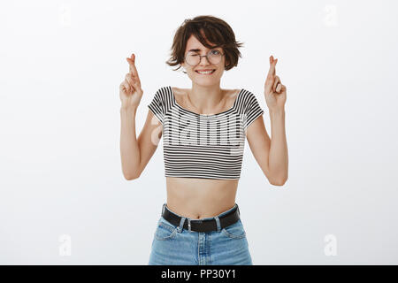 Praying for good result of interviewing. Stylish positive woman in round glasses ands stylish cropped top, smiling and biting lip peeking with one eye while crossing fingers and hoping, making wish Stock Photo
