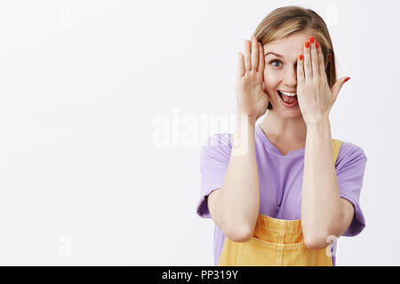 Horizontal shot of beautiful charming and playful female with fair short haircut, covering half of face joyfully, smiling broadly and playing peek-a-boo, having fun with siblings over gray background Stock Photo