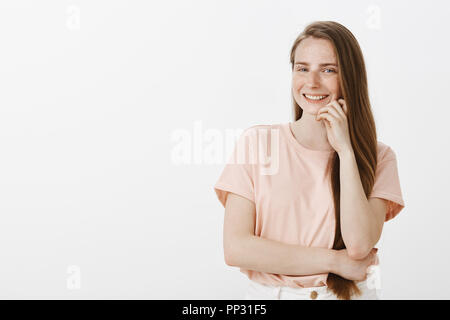 Waist-up shot of charming female freelance journalist with cute freckles and long brown hair, leaning head on hands and gazing at camera with amused carefree smile, standing over gray background Stock Photo