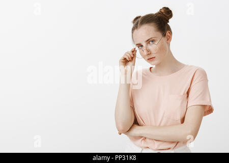 Cool confident trendy girl in stylish outfit, taking off glasses and staring self-assured and arrogant at camera, feeling like super star, standing over gray wall with hand crossed on chest Stock Photo
