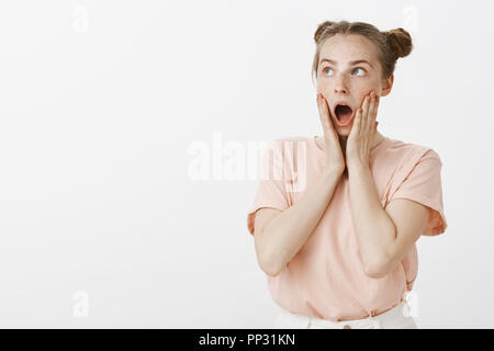 Girl being shocked seeing flying superhero in air. Amazed and speechless cute and funny woman with buns hairstyle, dropping jaw holding palms on cheeks, gazing at upper left corner astonished Stock Photo