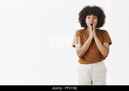 Indoor shot of impressed speechless good-looking african woman with curly haircut holding palms on face dropping jaw in amazement standing with opened mouth excited over gray wall Stock Photo