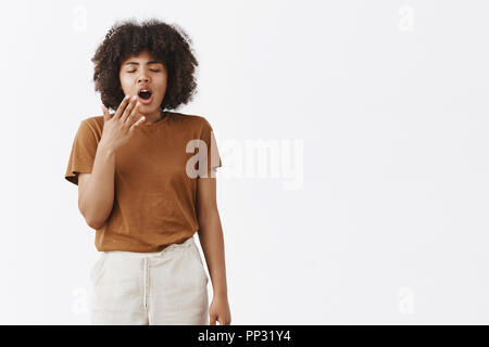 Hate jetlag. Portrait of sleepy cute African American woman with afro hairstyle closing eyes and yawning with wide opened mouth covering it with hand wanting sleep being tired over gray wall Stock Photo