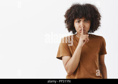 Waist-up shot of serious-looking displeased bossy african american girl in brown t-shirt frowning saying shush showing shh gesture with index finger over mouth demanding keep silent or save secret Stock Photo