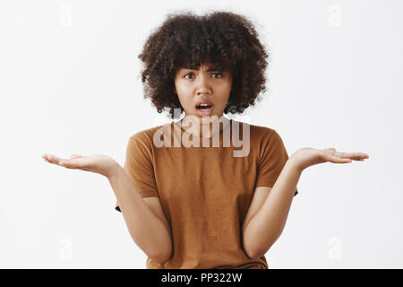 Waist-up sot of confused intense and annoyed african american young woman with curly hair in trendy brown t-shirt shrugging with palms spread aside making unaware and questioned look with opened mouth Stock Photo