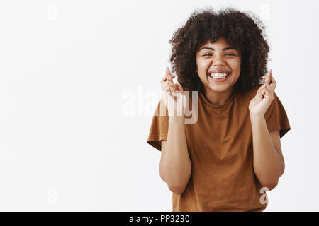 Waist-up shot of good-looking faithful and optimistic teenage african american girl with afro hairstyle crossing fingers for good luck and smiling joyfully praying for dream come true or making wish Stock Photo