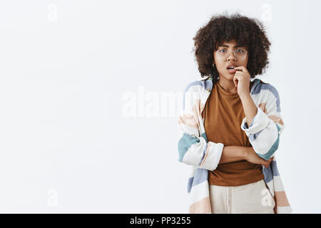 Waist-up shot of curious thoughtful and smart worried african american woman with afro hairstyle in transparent glasses biting fingernail and frowning while thinking how make hard choice Stock Photo