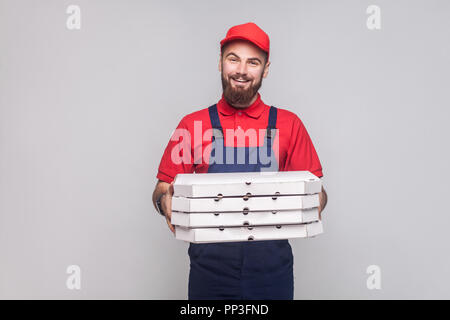 Young smiling logistic delivery man with beard in blue uniform and red t-shirt standing and holding stack of cardboard pizza boxes on grey background. Stock Photo