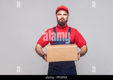 Portrait of young smiling logistic delivery man with beard in blue uniform and red t-shirt standing and holding the cardboard box on grey background.  Stock Photo