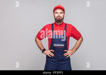 Portrait of young confident repairman with beard in blue overall, red t-shirt and cap standing and holding hands on waist with smile, indoor, studio s Stock Photo