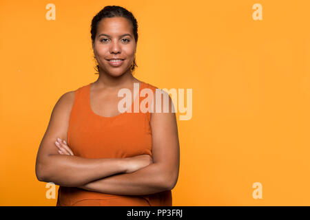 Young pretty mixed race black woman portait standing smiling with her arms crossed on an orange background Stock Photo