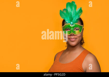 Young pretty mixed race black woman portait with green carnival mask on an orange background Stock Photo