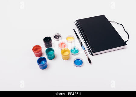 Set for drawing on white table: album, bright paints in open jars and one brush, top view. Stationery for hobby of painting. Stock Photo