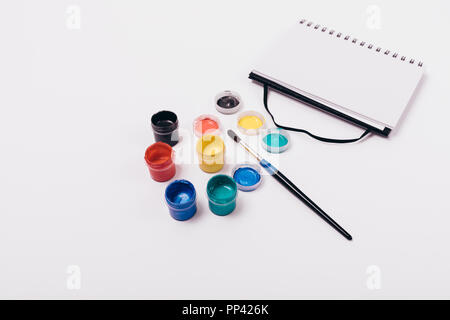 Opened notepad page near group of colorful paints in open jars, lids and single paintbrush on solid white background, top view. Stock Photo