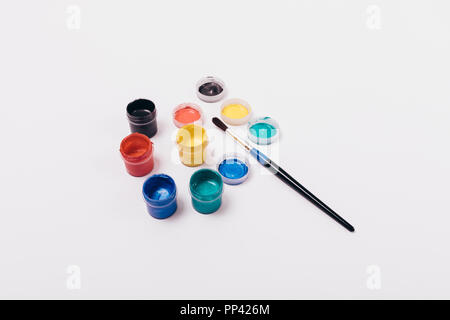 Group of multi-colored paints in open jars, lids and single paintbrush on solid white background, top view. Stock Photo