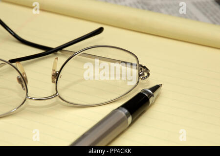 A yellow lined legal pad, reading glasses and a ballpoint pen in an office. Stock Photo