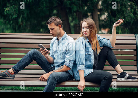 Phone addiction, young couple on the bench in park. Man and woman using their smartphones, addicted people Stock Photo