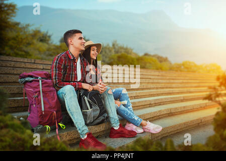 Couple of tourists with backpacks resting on the stone steps of ancient temple, excursion in town. Summer hiking. Hike adventure, travelling of young  Stock Photo