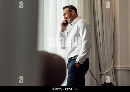 Businessman talking on phone while standing in a hotel room. CEO on business trip staying on a hotel and making phone call from landline phone. Stock Photo