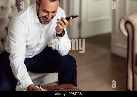 Mature businessman talking on phone and making notes. CEO of an organisation working from hotel room. Stock Photo