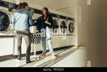 Smiling man and woman putting clothes in a washing machine together standing in a laundry room. Happy couple doing laundry together. Stock Photo