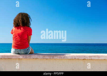 Mixed race African American girl teenager female young woman wearing a red t-shirt and denim shorts sitting on a wall looking out to a blue sea Stock Photo