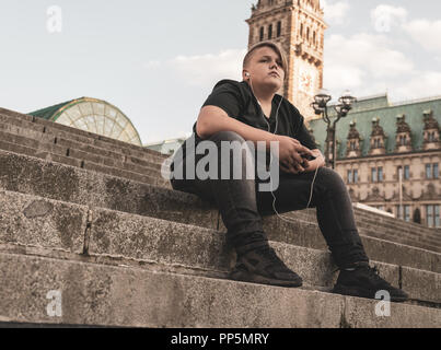 A young man wirh earphones is sitting on steps Stock Photo
