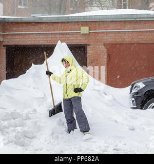 Cheerful girl with the shovel for snow removal stands near a huge snowdrift near the garage Stock Photo