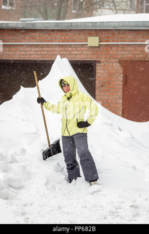 Girl with a snow shovel stands near a huge snowdrift during a blizzard Stock Photo