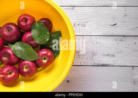 Wet ripe red apples with green leaves in bright round yellow plastic washbowl covered by water drops and placed on white old wooden table. Selective f Stock Photo