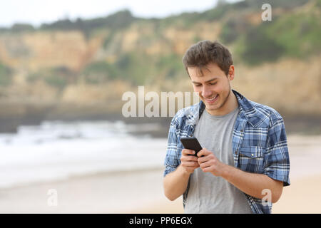 Happy teen guy using a smart phone walking on the beach Stock Photo