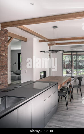 Trendy modern interior with white and brick walls and a ceiling with wooden beams. There is a kitchen island, table with chairs, chandelier, sofas, fi Stock Photo