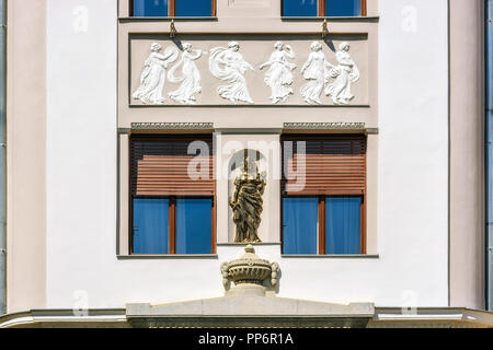 Two windows, half-closed with rolling shutters of brown color against a white wall with a gilded statue in a niche and a bas-relief from above. Stock Photo