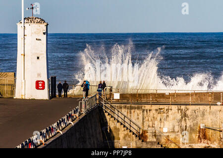 Waves breach the harbour wall and crash against the white lighthouse as crowds gather to watch on a sunny afternoon at Seahouses, Northumberland, UK. September 2018. Stock Photo