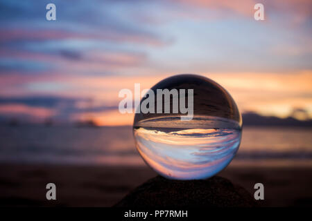 Sunset over Ocean with Reflection Captured in Glass Ball on Sand Stock Photo