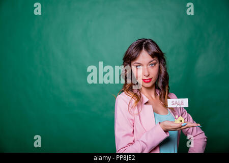 Beautiful smiling young woman in pink jaket with long hair, holding small wooden house in her hands with message for sale, isolated on green background, investment and payment concept. Copy space Stock Photo