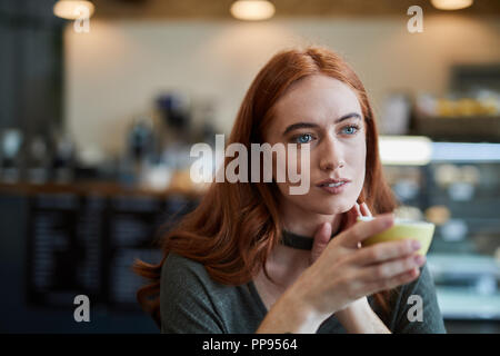 A single female, sits in a city cafe holding a hot beverage in a cup, looking thoughtfully off into the distance