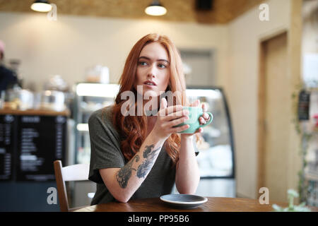 A female with an arm tattoo sits at a wooden table holding a cup of Cappuccino in a green cup Stock Photo