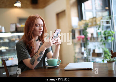 Red Head girl wIth arm Tattoo, sitting alone in a cafe looking at her mobile phone, with a laptop sat on the wooden table, Liverpool, UK Stock Photo