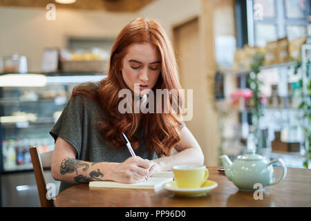 A female with shoulder length red hair sits in a city cafe at a wooden table writing in a notebook as a mobile phone sits next to her Stock Photo