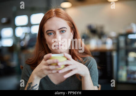 A single female, sits in a city cafe holding a hot beverage in a cup, looking at camera Stock Photo