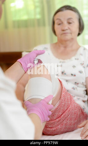 Doctor bandaging senior woman leg at home. Doctor giving first aid to patient. Focus is on doctor hand. Stock Photo
