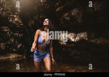 Young caucasian beautiful woman tourist in a waterfall pond in jungle looking up and smiling. Beautiful girl wearing bikini top and shorts in the pond Stock Photo